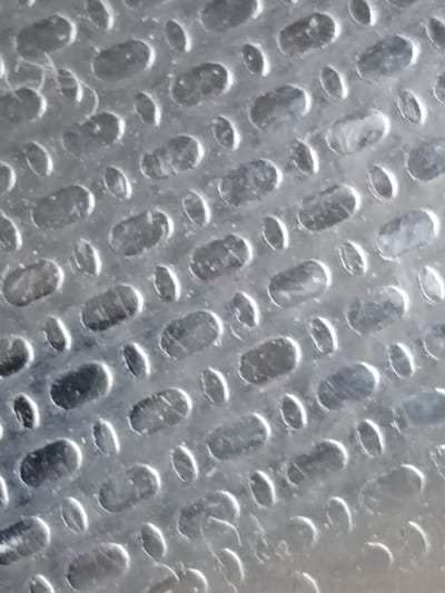 This is a close-up, abstract image of a textured metal surface. The surface has an embossed pattern of elongated oval indentations arranged in a staggered grid. The metal appears to be a dull, brushed silver or aluminum. The lighting creates highlights and shadows within the indentations, emphasizing the three-dimensional quality of the texture. There are no people, specific landmarks, or environmental details visible that would allow for localization in Temirtau, Kazakhstan or any other specific city. The image is purely focused on the texture of the material.