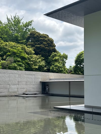 This image captures a view of the 21st Century Museum of Contemporary Art in Kanazawa, Japan. The scene features a modern architectural design, a reflecting pool, and dense natural foliage under a cloudy sky. A large, shallow reflecting pool occupies the lower half of the frame, its surface displaying ripples and reflections of the surrounding elements. To the far right, a tall, plain, light-colored building wall extends vertically, topped by a dark, flat, cantilevered roof overhang whose underside reveals a subtle grid pattern.
In the mid-left, a horizontal wall constructed from light grey, textured stone blocks provides a visual boundary. Set within this stone wall is a sheltered alcove or walkway, where a single individual dressed in light clothing stands, facing right. A minimalist bench or seating platform is positioned at the edge of the reflecting pool in front of the stone wall. Behind the architectural elements and dominating the upper left and central background, a dense expanse of lush green trees, displaying various shades from light to dark green, creates a vibrant natural backdrop. The sky above is overcast with soft, grey clouds.