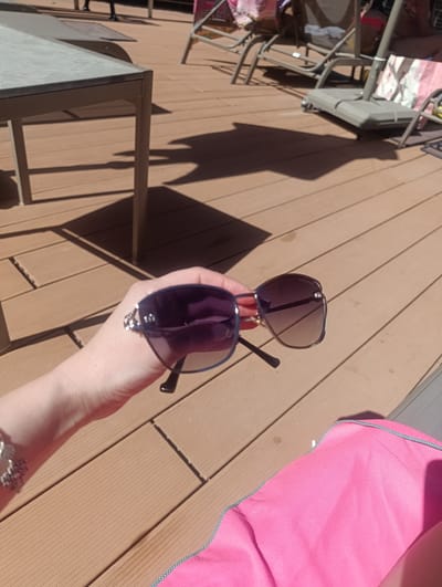 A close-up shot shows a person's hand holding a pair of sunglasses with purple lenses and a blue frame. The sunglasses are partially in focus, with the hand and a portion of a bracelet visible on the left side of the frame. The background is a wooden deck, with shadows cast by unseen objects, suggesting it is daytime. In the background, lounge chairs and a table are visible, indicating an outdoor setting, possibly a patio or pool area. A bright pink fabric, possibly a towel or beach bag, is in the lower right corner. The scene appears relaxed and leisurely, consistent with a sunny day outdoors. The location context is Urla, Turkey.