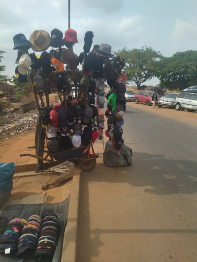 A street vendor is selling hats in Maraba, Nigeria, at the coordinates (latitude: 9.024, longitude: 7.573). The vendor has arranged a variety of hats, including baseball caps, fedoras, and bucket hats, on a wheelbarrow. There are over 30 hats displayed on the cart. The hats are arranged on sticks that are mounted in the wheelbarrow. The vendor is standing by the cart and talking to a potential customer. There is a large bag on the ground next to the cart. The scene is on a paved road with a dirt shoulder. There is a small group of cars parked on the side of the road.  One person is walking towards the vendor, and another is walking away. There is a broom leaning against the curb. There are piles of dirt and debris on the shoulder of the road. The sky is cloudy. The vendor is selling the hats in a busy street, with people walking by and cars passing.