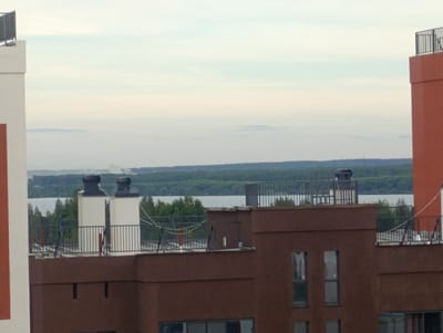 The image shows a view of urban rooftops and a distant body of water, possibly a river or lake, bordered by a tree-lined shore. The foreground features the tops of several modern buildings with flat roofs. One prominent structure on the left is partially visible, displaying a white facade and an orange accent. To its right, a brown building with multiple windows is seen. On the rooftops, there are various architectural elements including ventilation systems, railings, and what appear to be small structures or chimneys.

In the middle ground, another building section with a white structure and two dark, cylindrical ventilation units is visible, along with metal railings. Beyond this, the water stretches across the frame, reflecting a pale, overcast sky. A dense green forest occupies the far bank of the water. In the far distance, hazy outlines of more distant structures or terrain are faintly visible under the diffused light. The overall impression is an urban landscape viewed from an elevated perspective, with a focus on architectural features and the natural elements of water and forest. The scene is set in Zapadny, Russia, suggesting a contemporary urban development adjacent to natural scenery. No people are visible in the image.