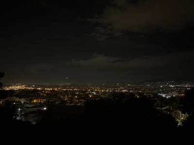 This image captures an elevated, panoramic nighttime view of the Gerakas Municipal Unit, Greece. The upper two-thirds of the frame are dominated by a dark, cloudy sky, with some faint light sources, possibly distant stars or aircraft, visible through thinner cloud patches. Below the horizon, the expansive urban landscape of Gerakas is illuminated by a dense array of artificial lights, predominantly warm yellow and cool white, delineating buildings, streets, and infrastructure. The lights extend across the middle ground and into the background, where more diffuse clusters of light indicate increasing distance. Hilly or mountainous terrain, also featuring scattered lights, is discernible in the far distance on the right. The immediate foreground and lower edges of the image are dark, consistent with unlit foliage or terrain from an elevated viewpoint. No discernible individuals, specific actions, or close-up objects are present within the frame; the scene depicts the static illuminated environment.