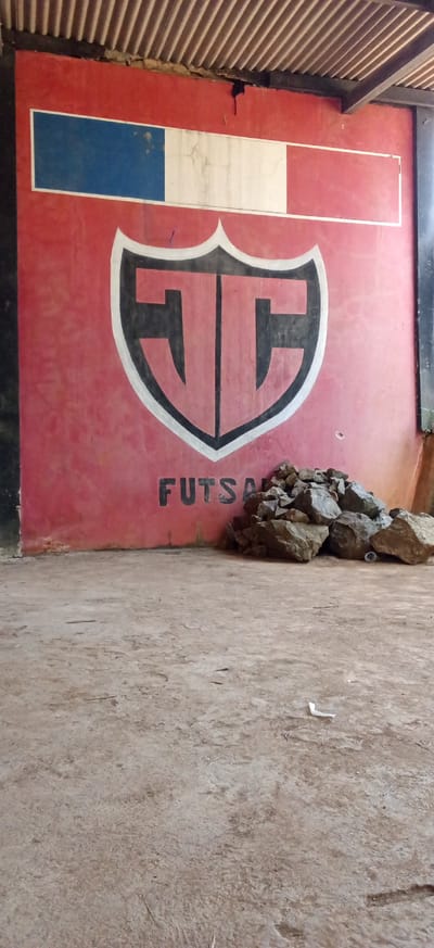 The image depicts a red wall with a painted emblem and the word "FUTSAL" below it. The emblem is shield-shaped with a black outline and contains two stylized black letters, possibly "T" and "C", on a lighter background. Above the emblem, a horizontal rectangle is painted with three vertical stripes: blue on the left, white in the middle, and white on the right. To the right of the word "FUTSAL", a pile of rocks is visible against the wall. The wall appears to be part of an outdoor sports facility. The ground in front of the wall is a dirt or concrete surface, with some debris scattered on it. The overall scene suggests an undeveloped or rustic sports area. The location is identified as the city of Ciapus, Indonesia.