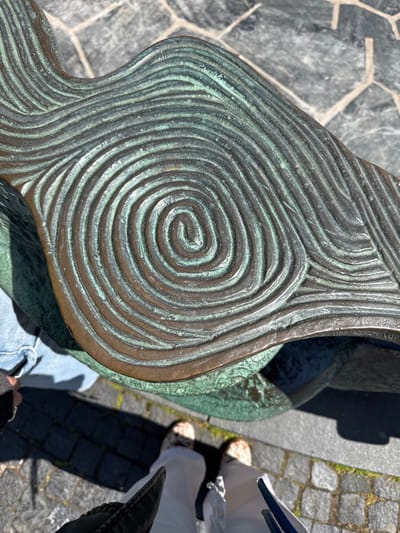 A high-angle shot captures a close-up of a bronze sculpture with a prominent spiral design, likely located in Malmö, Sweden. The sculpture exhibits a green patina, indicating oxidation. The background features a gray stone surface, possibly pavement, and parts of a person wearing jeans and another person in white clothing and neutral-toned shoes. A piece of dark clothing is also visible in the lower right of the frame.