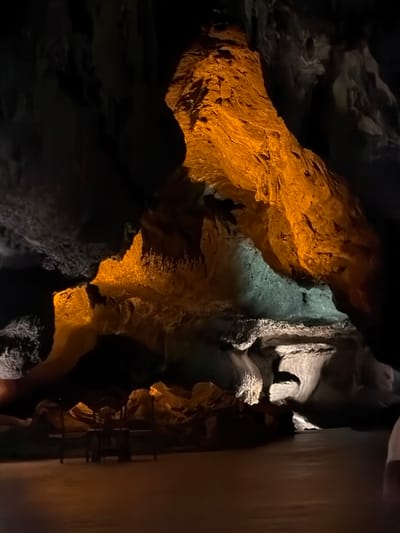 The image captures the dramatic, artificially lit interior of a large cave or grotto, set in Haría, Spain. The scene is dominated by rugged, natural rock formations displaying diverse textures and contours.

Strategic artificial lighting plays a crucial role in highlighting the cave's features and creating a high-contrast environment. A prominent, massive rock face on the left and central parts of the image is bathed in a warm, orange or amber light, emphasizing its rough, layered surfaces. In the middle of the frame, a body of water, likely a pool or subterranean river, reflects a cooler, greenish-blue light, contrasting sharply with the warm tones of the adjacent rocks. Other rock formations, particularly towards the right, appear in various shades of grey, with some areas receding into deep, dark shadows.

Evidence of human infrastructure for visitor access is visible on the far right. A smooth, light-colored, vertical wall or panel runs along the edge of the frame, suggesting a constructed walkway or barrier. Adjacent to this, a dark, ladder-like structure or safety railing can be discerned, indicating a prepared path for exploration.

The absence of natural light, combined with the artificial illumination, creates an atmosphere of mystery and awe, typical of a managed subterranean tourist attraction. No people are visible, and there is no discernible text within the image. The time of day and weather are irrelevant to this indoor scene.