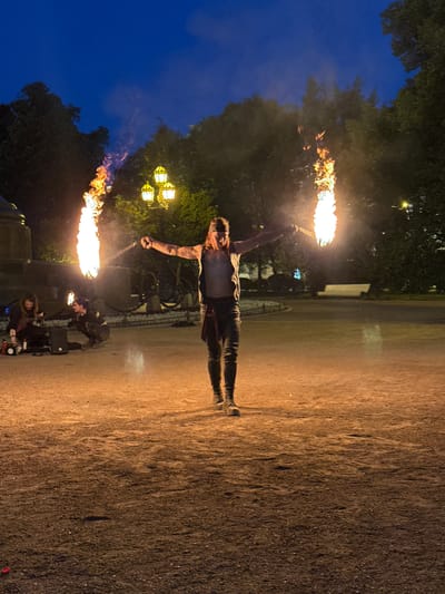The image depicts an outdoor performance at dusk, likely in a park or public square in Saint Petersburg, Russia. A performer is central to the scene, holding two flaming torches, one in each outstretched hand, creating arcs of fire against the darkening sky. The performer wears a dark sleeveless vest, dark pants, and a band around their forehead.
In the background, two other individuals are seated on the ground, appearing to be involved in the performance, possibly as support or fellow performers. One individual is playing a guitar, and near them are audio equipment, including a speaker. The environment features trees, a streetlamp with multiple glowing lights, and a park bench in the distance. The ground is covered in gravel or dirt, illuminated by the ambient light and the fire. The overall atmosphere suggests a public entertainment event taking place in a city park during the evening hours.