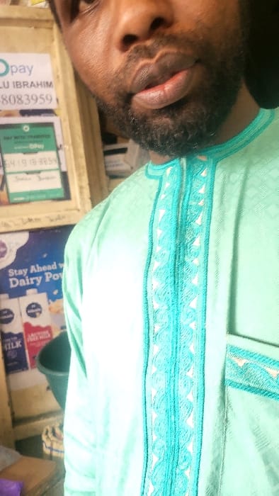 A dark-skinned male is depicted indoors, likely within a retail establishment in Fagge D1, Nigeria. The subject's face is visible from the upper chest to just above the eyes, featuring a dark beard and mustache. His lips are full and slightly parted. He wears a light mint green garment, possibly a traditional tunic, with intricate turquoise and white embroidery on the central placket and a small design on a visible pocket. The fabric of the garment exhibits a subtle patterned texture. In the background on the left, a wooden surface serves as a display for multiple signs. A white Opay service sign at the top displays "LU IBRAHIM" and a partial phone number "8083959". Below this, another Opay sign with "PAY WITH CHARGES" and a partial number "6419183854" is visible. Further down, a poster advertises "Dairy Pow" with depictions of milk cartons. A dark green bucket and a folded white cloth with colored stripes are partially visible at the bottom left. The background to the subject's right is dark and indistinct.