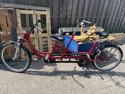 A red tandem cargo bicycle is parked outdoors on a paved surface, casting a shadow. The bicycle has two seats, handlebars for both riders, and a blue cargo box located between the seats. Behind the bicycle, a wooden fence and stacked wooden pallets are visible. The environment appears to be in Lisse, Netherlands.
