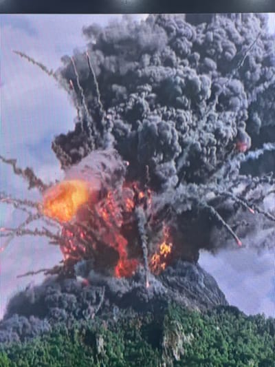 The image depicts a massive explosive volcanic eruption occurring in Apshak, Uzbekistan. A colossal, dense column of dark grey ash and smoke billows upwards, occupying the majority of the frame's upper section. Within this turbulent plume, numerous bursts of bright orange and red incandescent material are visible, indicating intense heat and explosive activity. Multiple glowing projectiles, appearing as streaks of fire and smoke, are ejected radially from the eruption column, arcing against the lighter sky background on the left. The lower portion of the image reveals the volcanic mountain's slopes, covered densely with green foliage. The sky on the left and top-left is a clear light blue with some faint cloud formations. No individuals or man-made structures are discernible within the visible scene.
