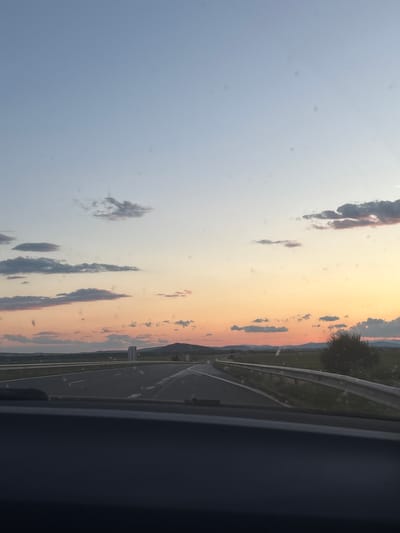 A view from a vehicle dashboard shows a highway in Zimnitsa, Bulgaria at sunset. The sky is a gradient of pale blue at the top transitioning to orange and pink hues near the horizon, with scattered clouds. The highway, with lane markings visible, curves to the left in the distance. A metal guardrail runs along the right side of the road. Beyond the highway, a grassy field and distant hills are visible. The dashboard of the vehicle is dark and takes up the bottom portion of the frame.