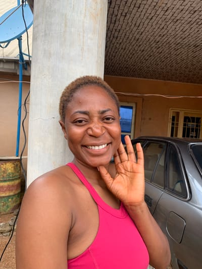 A medium shot captures a Black woman with short, light brown hair, fair skin, and a warm smile, posing outdoors. She is wearing a bright pink tank top. Her right hand is raised, palm facing the camera, in a waving gesture. Behind her, a weathered, pale grey concrete column stands to her left. To the right of the column, a silver car is parked. Further in the background, a building with brown walls and windows is visible, with a textured, brick-like ceiling overhead. A blue satellite dish is mounted on a pole to the far left, and two yellow and green barrels are stacked near the base of the column. The overall setting appears to be a residential or commercial area in Russo, Nigeria, characterized by its utilitarian architecture and outdoor elements.