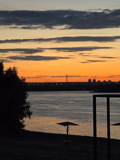 The image depicts a serene sunset over the Volga River in Zhigulyovsk, Russia. The sky is a vibrant gradient of orange and yellow, with dark gray clouds streaking across the upper portion. The river's surface reflects the warm hues of the sky, creating shimmering patterns.

On the left bank of the river, a silhouette of a dense cluster of trees stands out against the colorful sky. The right bank is lined with a dark silhouette of buildings and a tall radio tower, suggesting an urban presence. In the foreground, the dark silhouette of a beach is visible, with a few beach umbrellas and a portion of a dark, rectangular structure. The overall scene conveys a sense of tranquility and the peaceful end of a day.