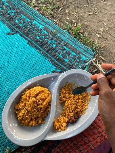 A person's hand holds a black spoon over a gray plate divided into two sections. The top section contains a pile of rice and a piece of meat, while the bottom section holds a mound of rice. The plate sits on a woven mat, with blue and black sections on the left and brown and red sections on the right. Grass and dirt are visible in the background near the blue and black part of the mat.