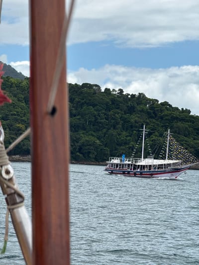 The image depicts a maritime scene in Angra dos Reis, Brazil, under a partly cloudy sky. In the foreground, a blurred reddish-brown wooden vertical beam, identified as part of a vessel's mast or rigging, occupies the left side of the frame, showing ropes and metal fixtures. A small red fabric detail is visible at the extreme upper left. In the midground, a white tour boat with red and dark blue trim, featuring two decks (one enclosed, one open) and two masts, is underway, moving from right to left across rippled grey-blue water. The masts are adorned with numerous small yellow triangular pennants. Multiple indistinct human figures, presumed to be passengers, are visible on both decks of the tour boat. The background features a densely forested, tropical green mountain or island coastline bordering the water. The sky above displays a mix of white cumulus clouds and segments of clear blue.