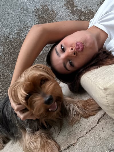 A young adult female and a Yorkshire Terrier dog are lying on a cracked concrete surface in Galisteo, Spain. The female, positioned diagonally with her head tilted, has dark hair, wears a white t-shirt, and purses her lips into a kissing expression while looking directly at the camera. Her left hand is placed on the dog's neck/chest area. The Yorkshire Terrier, with brown and black fur, lies adjacent to her, its mouth slightly open, revealing small teeth. The background consists of a textured wall, possibly plastered, exhibiting varying shades of grey and brown. A light-colored fabric item is partially visible near the female's right shoulder.