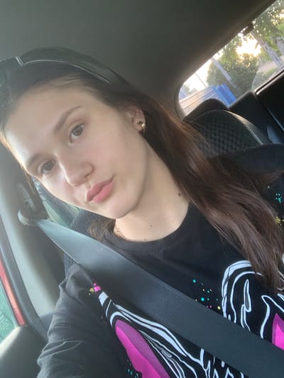 A young woman with long, dark brown hair is seated in the passenger seat of a car, facing forward, and looking at the camera. She is wearing a black t-shirt with a graphic design featuring pink and white shapes and colorful dots. A grey seatbelt is fastened across her chest. The interior of the car is visible, including the headrest of the seat behind her. Sunlight filters in through the side window, illuminating trees and a blue structure outside. The image appears to have been taken in Belogorsk, Russia, based on the provided context.