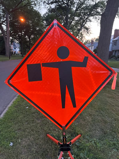 The image shows a diamond-shaped orange construction sign depicting a silhouette of a person holding a flag. The sign is on a tripod stand placed on a grassy area beside a paved sidewalk and road. In the background, there are trees, houses, and a street lamp. The sky is overcast, suggesting it is either dawn or dusk. The sign itself has reflective properties, indicated by the textured surface and the way it catches the light. Text on the sign reads "FLAGGER SYMBOL". The setting appears to be a residential street in Shaker Heights, Ohio, United States of America.