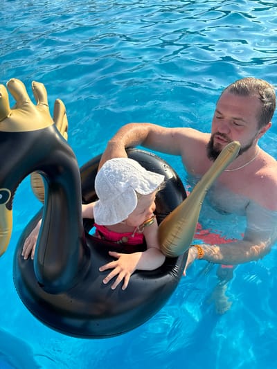 A man and a baby are in a swimming pool. The man is partially submerged in the water and is holding onto an inflatable swan. The baby is sitting inside the inflatable swan and is wearing a white hat and a pink swimsuit. The water is clear blue, and the sunlight is reflecting off the surface. The scene takes place in a swimming pool. The city of Колокша, Russia is mentioned as a location, but there is no visual information in the image to confirm or deny this.