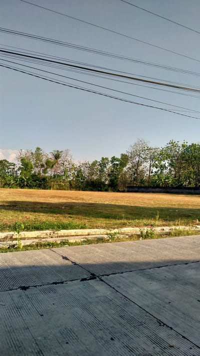 This media file is a landscape photograph taken outdoors, likely in Nganjungganjung, Indonesia. The scene depicts a dry, yellowish field, possibly a sports field, with a yellow soccer goal visible in the distance. A line of trees forms a backdrop to the field, with some taller trees and foliage suggesting a tropical or subtropical environment. A concrete sidewalk with distinct grooved lines runs across the foreground, casting a shadow. Overhead, multiple power lines stretch across the clear, pale blue sky.
The time of day appears to be late afternoon or early morning, indicated by the long shadows cast on the sidewalk. The weather seems clear and sunny, with no visible clouds. The dry state of the field might suggest a dry season. The overall mood is tranquil and somewhat arid, with a sense of open space. There are no people visible in the image, and no text is present. The focus is on the natural landscape and man-made elements like the field and power lines.