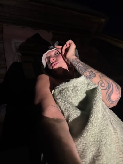 A woman with blonde hair, wearing a gray headband and wrapped in a pale green towel, is lying down. Her right arm, adorned with tattoos, is visible. Her eyes are closed, and her right hand is near her face, touching her forehead. The background appears to be a dark, rustic attic space with wooden beams and paneling, consistent with a location in Maksimovo, Russia. The lighting is dim, casting deep shadows.
