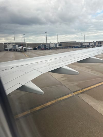 The image shows the wing of an airplane at Cleveland Hopkins International Airport in Cleveland, Ohio, United States of America. The aircraft is parked at a gate. Airport infrastructure is visible, including ground support equipment, jet bridges, and other aircraft parked nearby. The tarmac is wet, and the sky is overcast.