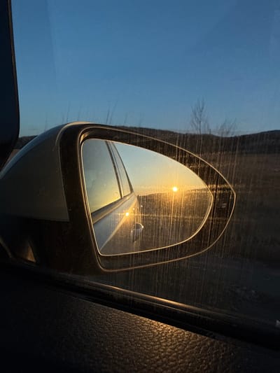 The image displays a rural landscape at either sunset or sunrise, observed from the interior of a vehicle in Relyovo, Bulgaria. The lower right portion of the frame shows the dark, textured surface of a car's interior door panel. A grey side mirror housing is visible on the left, enclosing the side mirror itself. The mirror's surface reflects a prominent, bright orange-yellow sun positioned low on the horizon, casting a strong glare and illuminating a flat, open area, possibly a road or field, behind the vehicle. Streaks and dust are visible on the glass of both the car window and the side mirror. Beyond the mirror, through the car window, a dry, uncultivated terrain with sparse, bare vegetation or tall grasses stretches towards a distant, dark horizon line, indicative of hills or mountains. The sky above is clear and light blue, transitioning from brighter hues near the horizon. No individuals are visible in the scene.