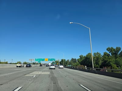 The image shows a clear blue sky above a multi-lane highway in Sacramento, California, United States of America. The highway is filled with various vehicles, including cars, a pickup truck, and a trailer, all traveling in the same direction. Overhead signs indicate directions to "WEST 50 San Francisco" and "SOUTH 44 Los Angeles." Trees and vegetation line the right side of the highway, with a concrete barrier separating the lanes from the embankment. A street light stands tall on the right side of the road, its arm extending over the lanes. The road surface shows wear and tear, with some patches and markings visible. The scene suggests a typical sunny day on a major freeway.