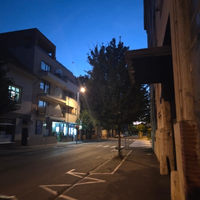 This is a nighttime street view in Piešťany, Slovakia, featuring a multi-story building on the left, a tree-lined street, and a building on the right. The sky is a deep twilight blue. The building on the left is modern, with balconies and large windows, some of which are illuminated. A streetlamp casts a warm glow on the street and sidewalk. On the right, a building with a textured facade and exposed brick is visible, partially obscuring the view further down the street. The street itself has white lane markings and a pedestrian crossing. There are no people or vehicles prominently featured in the scene. The overall impression is of a quiet urban street after dusk.