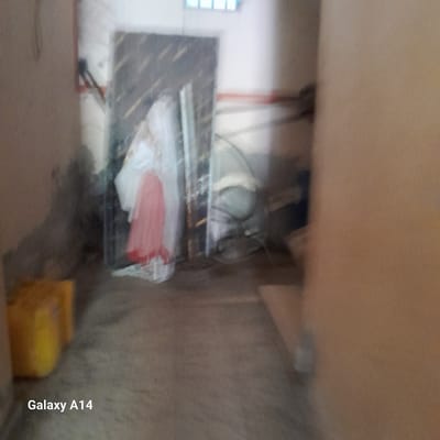 The image shows an indoor scene. In the foreground on the left, there are two yellow barrels. In the middle ground, there is a large, semi-transparent sheet leaning against a wall, with a pink and white fabric draped over it. Behind this, there is a large, dark, possibly metal object. The walls are a light color, and there are some horizontal lines that may be pipes or conduits on the wall to the right. The floor appears to be concrete. There is a watermark in the bottom left corner that says "Galaxy A14". The image is blurry and lacks detail, making it difficult to ascertain the precise nature of the objects or the environment. There are no people visible in the image. Based on the watermark, it is likely a photograph taken with a Samsung Galaxy A14 phone. The location is stated as Deir el-Balah, Palestinian Territories, but this information cannot be independently verified from the image itself.