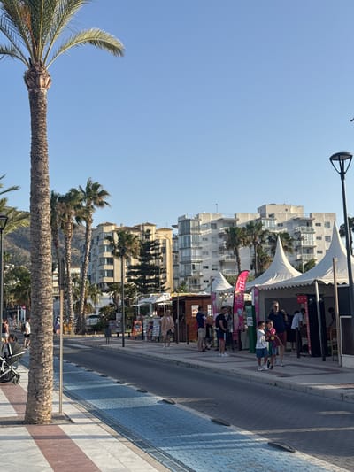 In Pla Parcial Platja, Spain, a paved street runs alongside a sidewalk lined with palm trees. Several buildings, including multi-story apartment complexes, are visible in the background. On the sidewalk, people are walking and standing, some near tents that appear to be part of an outdoor market or event. A lamppost stands on the right side of the image. The sky is clear and blue.