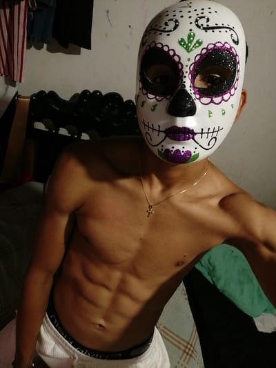 A shirtless person wearing a decorative sugar skull mask poses in a selfie. The mask is white with colorful floral designs around the eyes and mouth, and black outlining. The person is wearing a gold necklace with a cross pendant and white shorts with a black waistband. The background includes a bed with a green pillow, a black headboard, and hanging clothes.
