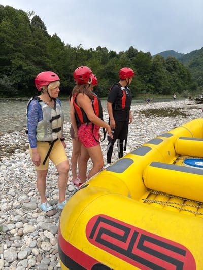 This media file depicts four people standing on a rocky riverbank next to a bright yellow raft. All individuals are wearing red helmets and life vests, suggesting they are preparing for or have just finished a rafting activity.

The subjects include two women in the foreground and two individuals behind them. The woman in the very front wears a blue patterned long-sleeved shirt and yellow shorts, with a grey and white life vest. Her blonde hair is short, and she appears to be looking up and to the left, with a neutral expression. Behind her, another woman, with brown hair, is wearing a red and black life vest over a pink patterned garment and dark shorts. Her head is tilted down. The two individuals furthest back are less visible, but one appears to be wearing black shorts and a black and red life vest, with a red helmet.

The scene is set by a river with a rocky bed, and a dense green forest covers the background hills. The sky is overcast, suggesting a cloudy or possibly rainy day. The raft itself is large, yellow, and appears to be made of durable material, with black and red accents and a coiled rope attached to it. The rocky terrain of the riverbank is detailed, with a variety of grey and white stones.

No specific activity is clearly captured in this still image, but the attire and the raft strongly indicate a rafting excursion. The overall mood seems to be one of preparation or a pause during an outdoor adventure.

Visible text includes "aqua design" on the grey and white life vest in the foreground. The logo on the yellow raft, a red geometric design with black outlines, is also a notable detail. The location context provided is Džajići, Bosnia and Herzegovina.