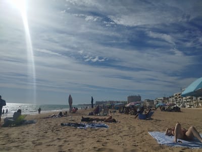 This is a wide shot of a sandy beach on a sunny day with a bright sunbeam cutting across the sky from the left. The sky is a vibrant blue with wispy white clouds. The beach is populated with many people, some sunbathing on towels and lounge chairs, others walking or standing. The ocean is visible in the background, with gentle waves and a few people in the water. Buildings line the horizon behind the beach, suggesting a coastal town. Several beach umbrellas and chairs are scattered across the sand, some occupied and some empty. The overall atmosphere appears relaxed and leisurely, with people enjoying a typical beach day. The lighting suggests it is midday. There is no visible text in the image.