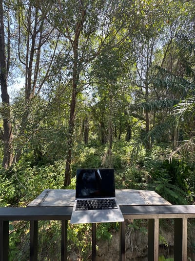An open silver laptop, consistent with a MacBook model, is positioned on a weathered wooden surface in an elevated outdoor setting in Phra Ae, Thailand. The laptop's dark screen displays ambient reflections, and its keyboard is visible. The wooden surface, which appears to be a table or shelf, is situated above a dark metal railing composed of vertical and horizontal bars. The laptop's front edge extends slightly beyond the wooden surface and over the railing. The background consists of a dense, lush tropical forest under bright daylight conditions, characterized by numerous tall trees with vibrant green foliage and sunlight filtering through the canopy. No individuals are present in the scene.