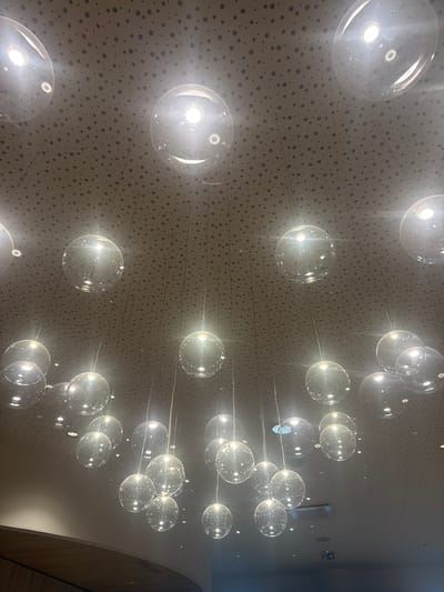 The image shows a ceiling with a decorative lighting installation. The ceiling is light-colored with a pattern of small, dark circles. Suspended from the ceiling are numerous clear glass or plastic spheres, each illuminated from within, creating bright points of light. Thin wires or rods connect the spheres to the ceiling. The spheres are arranged in clusters, seemingly at varying heights. To the right side of the frame, a portion of a curved wall is visible. Based on the prompt mentioning Marcq-en-Barœul, France, this is likely a feature inside a building in that city. There are no people visible in the image.