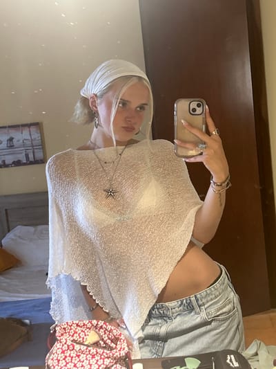 The media file is a portrait of a young woman taken in a room, reflected in a mirror. The woman is wearing a white, sheer, loose-fitting top, denim shorts, and a white headscarf. She has blonde hair and is wearing several rings and a necklace with a star-shaped pendant. She is holding a smartphone in her right hand, taking the photo of herself in the mirror. The background shows a wooden door and a wall with a framed picture. A red and white floral bag is visible on the right side of the frame. The lighting suggests it is daytime. The overall mood appears casual and relaxed. No visible text is present in the image.