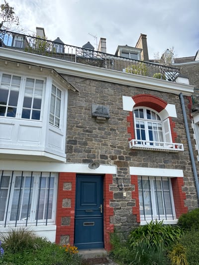 The image shows the facade of a stone building located in Dinard, France, at the coordinates 48.631 latitude and -2.052 longitude. The building has a bay window on the left, a blue door in the middle, and another window on the right.  A white iron railing runs across the front of the house, and there is a plaque above the door with a sailboat image on it. There is a white railing in front of the window on the right. The building has a stone facade with red brick accents. There is some vegetation in front of the building. The sky is cloudy.