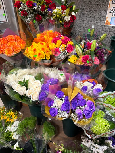 This is a medium shot photograph of a vibrant display of assorted flower bouquets. The flowers are arranged in multiple clear plastic wrappers and are presented at various heights and angles, suggesting they are for sale.

In the foreground, on the left, a bouquet of yellow freesia and white flowers is visible. To its right, two bouquets of bright green, fluffy flowers, possibly carnations or similar, are displayed in black plastic buckets. Further to the right, a bouquet of light green hydrangeas wrapped in clear plastic is prominent.

In the middle ground, a large bouquet of white roses is centered, with a bouquet of purple roses to its right, both wrapped in clear plastic with some colorful paper accents. Behind these, a bouquet of mixed yellow, orange, and pink roses and daisies is visible. At the top, a bouquet of red roses is featured.

The background consists of a textured, grey, speckled wall, reminiscent of granite or stone, which appears to be the interior of a store or market. A wooden element is visible on the left edge of the frame, possibly part of a shelving unit or display. A sign with some text and graphics is partially visible on the far right background.

The lighting is bright and even, illuminating the colors and textures of the flowers. No people are present in the image. The overall impression is a well-stocked floral display, likely in a flower shop or a market stall within New York City, USA.