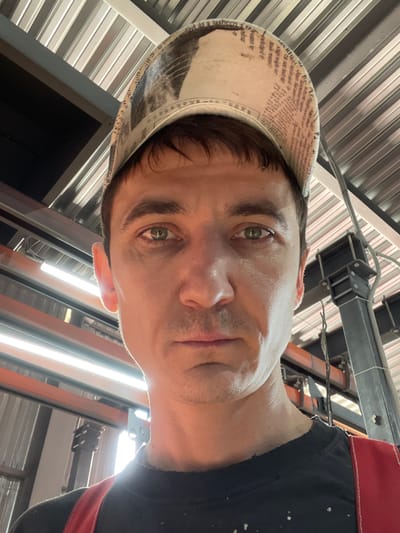 A man wearing a camouflage patterned baseball cap and a black t-shirt with red straps is pictured from the chest up, looking directly at the camera. He has short brown hair and a neutral expression. The background consists of industrial-style metal roofing and lighting fixtures, suggesting an indoor workspace. The lighting is bright, casting some shadows on the man's face. This image was taken in Сокуры, Russia.