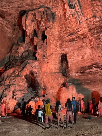 The image depicts a cave-like structure with rough, textured walls, illuminated by red lighting, in Gollagutta, India. The cavernous interior features natural rock formations, including stalactites and crevices. In the foreground, a group of individuals are gathered. Some appear to be dressed in traditional or ceremonial attire, with draped fabrics and elaborate hairstyles. One person is seated on a white plastic chair. Further back, a wooden ladder leans against a rock face, leading into an upper recess of the cave. Several figures are positioned in the background, some standing and others appearing to be seated or engaged in activities within the cave. The ground is covered in dirt and loose stones. The scene suggests a cultural or religious gathering or performance taking place within this natural environment.