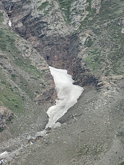 The image depicts a steep, rocky mountainside in the Gemur region of India. A large patch of snow is visible in a ravine, partially melted and forming a stream at its base. The surrounding terrain is rugged, with sparse vegetation and loose rocks. The image is taken from a distance, offering a wide view of the natural landscape. There are no people or man-made objects prominently visible in the scene.