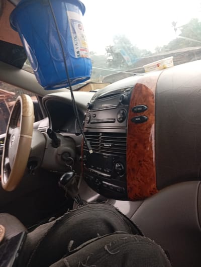 The image depicts the interior of a vehicle, likely a minivan, viewed from the passenger side. A blue plastic bucket hangs from the roof, partially obscuring the windshield. A portion of the dashboard is visible, featuring the steering wheel on the left, a gear shift, and the car's audio and climate control system. Wood-grain trim accents the central console. In the foreground, a person's legs, clad in ripped denim jeans, are visible, suggesting they are sitting in the driver's or passenger seat. The environment outside the vehicle appears to be an outdoor setting, possibly rural, with some greenery and what looks like rustic structures in the background. This scene is located in Imilike, Nigeria.