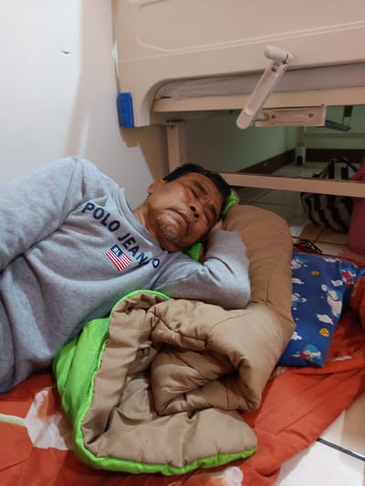 An adult male with dark hair and stubble is sleeping on his right side, facing the camera. He is wearing a gray long-sleeved shirt with "POLO JEANS CO" printed on it, along with an American flag patch. He is covered by a brown blanket and a green sleeping bag, resting his head on a tan pillow. Next to the pillow is a blue pillowcase with cartoon characters. The man appears to be in a room with a white wall and a white elevated structure with a crank mechanism, possibly a hospital bed or a bunk bed. The floor is tiled and there are some bags in the background. The scene is illuminated by indoor lighting. The location is described as being in the city of Karangtengah, Indonesia.