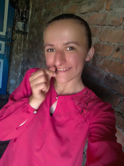 This is a selfie of a young woman with her hair pulled back and wearing a vibrant pink blouse. She is standing in front of a brick wall, with a partial view of a blue door and some framed pictures on the left side of the image. The woman is smiling and has her fingers near her mouth. The lighting suggests it is daytime. The scene appears to be outdoors or in an open-air structure, possibly a porch or an entryway, given the rustic brickwork and the presence of a door. The overall mood is casual and friendly. There is no visible text in the image.