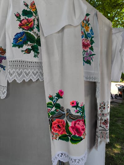 The image depicts multiple embroidered textiles, likely traditional Ukrainian rushnyky or clothing items, displayed outdoors in Illintsi, Ukraine. In the foreground, white and off-white fabric pieces with colorful, cross-stitched embroidery are vertically arranged. The leftmost visible textile features large floral motifs in red, blue, yellow, and green, with a scalloped white lace trim at its lower edge. Centrally positioned, a longer vertical textile displays two grey-brown birds nestled among prominent red and pink flowers with green foliage, also finished with a white lace border. To the right, a partially visible textile exhibits more intricate, geometric and floral patterns, including a stylized cross, in red, black, and other colors. The fabrics appear to be hanging from an unseen support structure. In the background, a light grey surface is present behind the textiles. Beyond this, green foliage from a tree canopy is visible at the top right, and green grass extends towards the lower right. A blurred dark object with wheels, possibly a utility cart, is partially visible on the grass in the far right background. The scene is illuminated by bright daylight. No individuals are present.