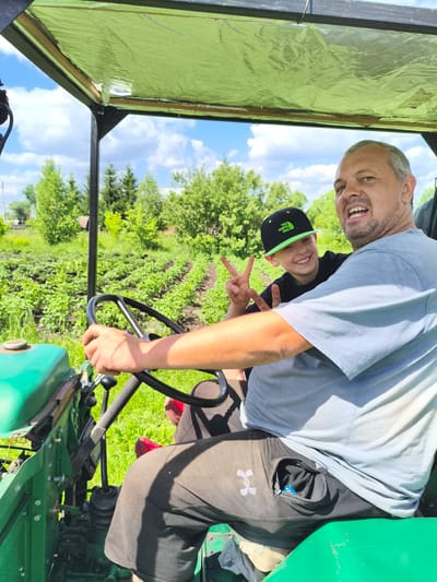 In the city of Ilmino, Russia, a man and a boy are sitting in a tractor. The man is driving, with his hands on the steering wheel. The boy is sitting next to him, smiling and making a peace sign with his fingers. They are both wearing casual clothes. The tractor is green and appears to be old. In the background, there is a field of green plants, likely a garden or farm, with trees and a blue sky with white clouds. The overall scene suggests a casual outing or work in a rural setting.