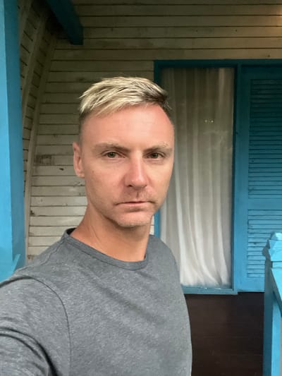 A fair-skinned man with blonde-tipped hair and a gray t-shirt faces the camera. He's positioned in front of what appears to be a light blue porch or veranda, with vertical wood paneling visible. A window with a white curtain and blue frame is behind him. The scene looks like an outdoor setting with natural light illuminating the subject.