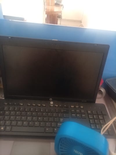 A black laptop computer is positioned on a desk, with its screen facing forward. The keyboard is visible below the screen, and a blue fan is situated in the foreground, partially obscuring the keyboard. In the background, a blurry office environment can be discerned. The image is taken from a slightly elevated angle, looking down at the laptop and fan. The scene appears to be an indoor setting, likely an office or a workspace. The location is Kinkinau, Nigeria.