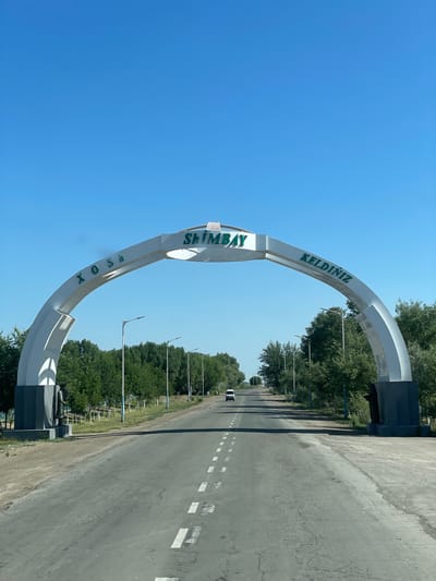 An archway over a two-lane asphalt road reads "XOSH SHIMBAY KELDINIZ" in green lettering. The archway is white with dark grey bases. On the left base of the archway is a dark statue of a standing figure. The road is lined with trees on both sides, and streetlights are visible. The sky is clear and blue. A white van is driving away from the camera in the distance. The location is the city of Ayteke, Uzbekistan.