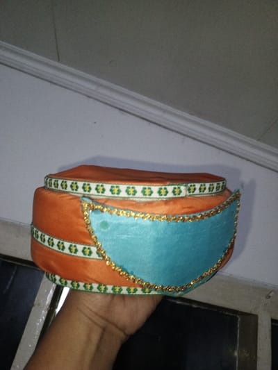 An individual, whose right hand is visible in the lower center, holds an intricately decorated cap or turban, likely for a ceremonial purpose, in an indoor setting in Na Sang, Laos. The cap features an orange, velvety main body wrapped horizontally with two parallel strips of white ribbon patterned with green, four-leaf clover-like motifs. A prominent, light blue, semicircular panel adorns the front of the cap, outlined by a continuous gold-colored, beaded trim. The blue panel exhibits two faint, darker blue-green smudges on its upper left quadrant. The background consists of a light gray wall with a white architectural molding running diagonally from the upper left to the upper right. To the lower right and behind the hand, a darker, possibly recessed area or window frame is visible. The lighting is consistent with an indoor environment.